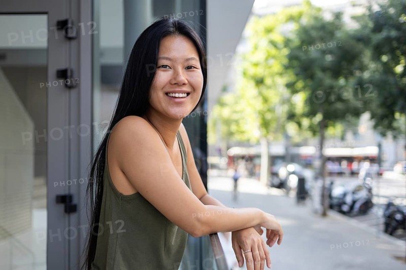 A Chinese woman smiles while leaning on a balcony in a bustling city. The sun shines brightly, capturing the essence of a vibrant summer day in an urban environment