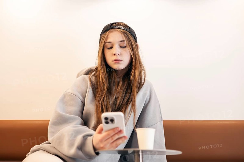A teenage girl in a casual hoodie and cap is intently using her smartphone while sitting on a modern brown sofa, with a cup on the table in front of her