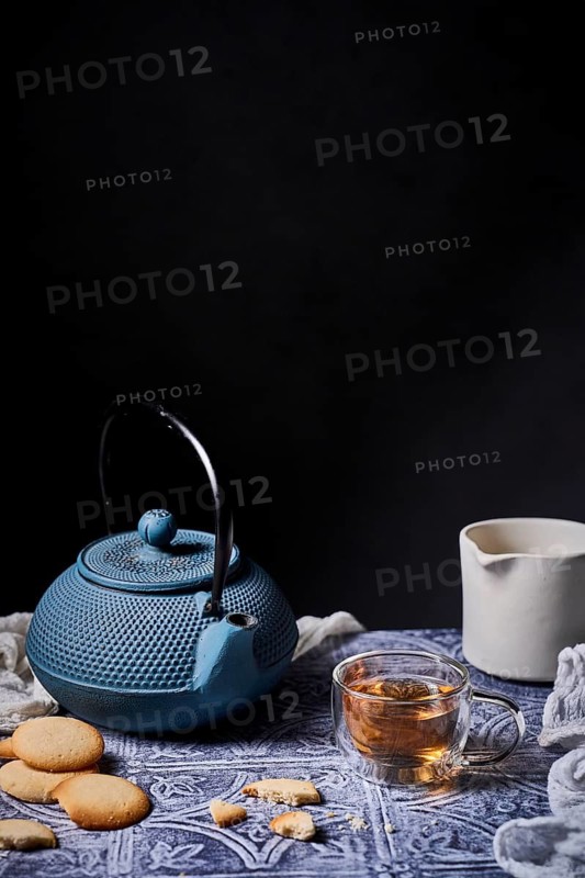 A cozy tea time setup featuring a blue teapot, a steaming cup of tea, and delicious cookies on a patterned cloth. A milk jug adds a touch of elegance to the scene