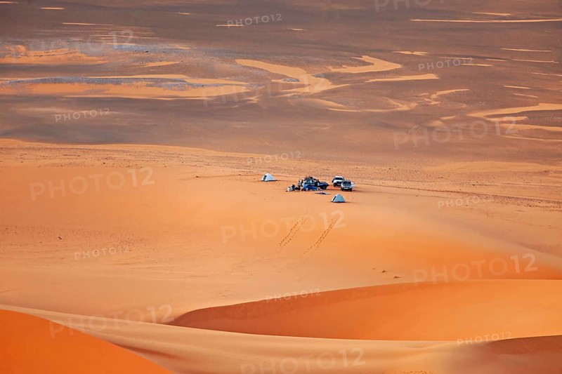Camp with cars and tents in a vast, tranquil sandy desert landscape under an orange-coloured sky, tent camp in the endless sandy landscape of the Sahara in Algeria