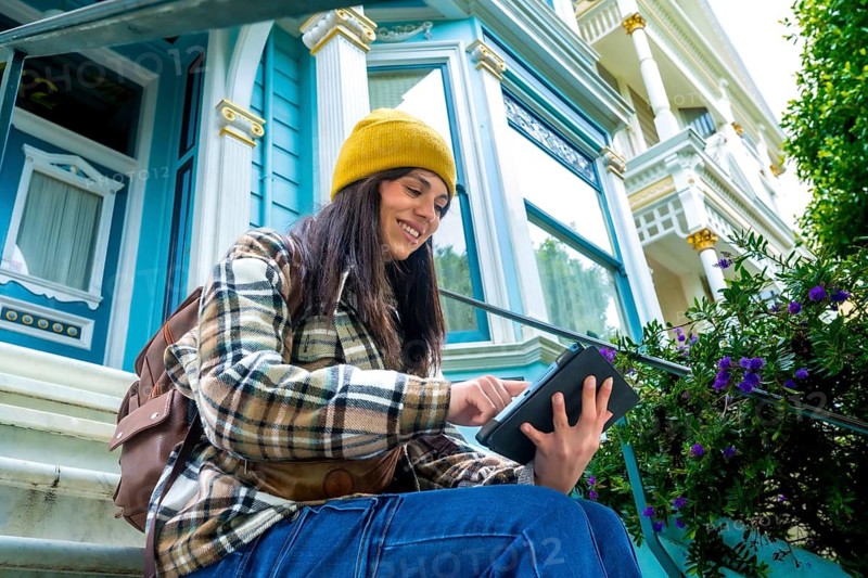 Woman wearing a beanie, plaid shirt, and jeans sitting on steps in front of a blue victorian house, smiling and interacting with a digital tablet on a city street