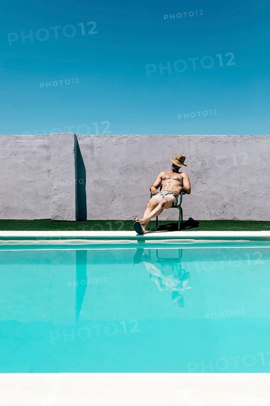 A man takes a nap relaxing on a chair by a swimming pool under a clear blue sky. He wears a straw hat and shorts, enjoying the sunny day, with a concrete wall behind him