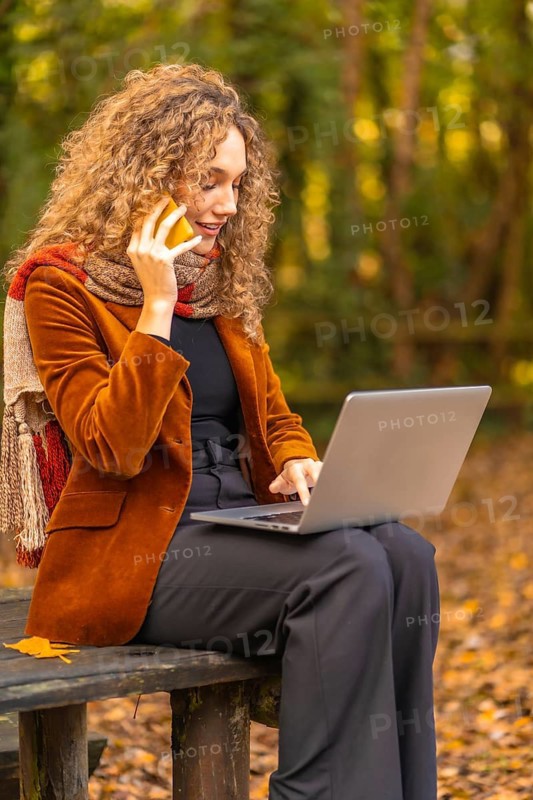 Vertical photo of a woman with curly hair using mobile while working with laptop in a park in autumn
