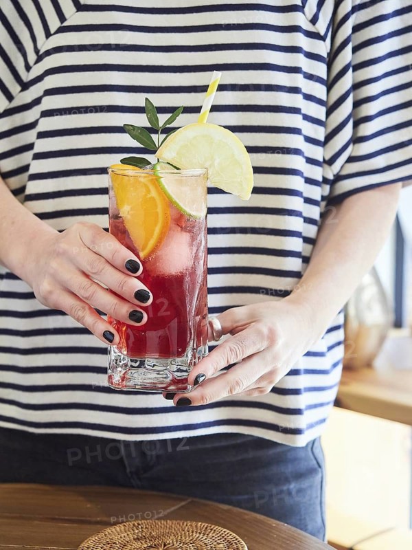 Refreshing cold red citrus drink with fresh lime, orange and lemon slices in glass in the hands of woman in striped sweater