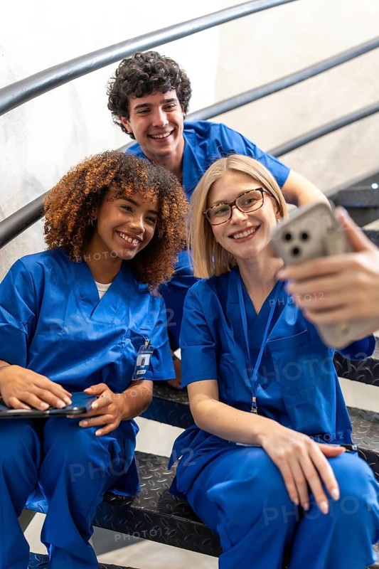 Group of smiling young healthcare professionals wearing blue scrubs sitting on steps, posing for a self portrait on a smartphone, representing education, teamwork, and friendship in medicine