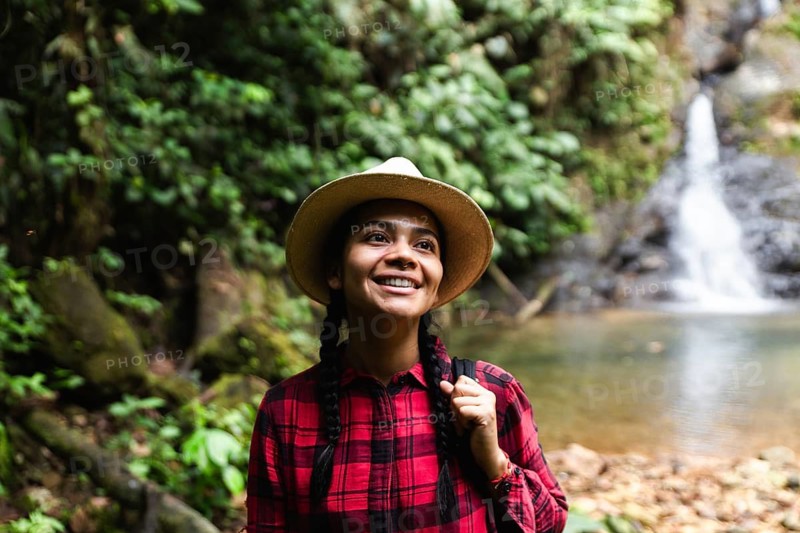 Portraying a joyful Ecuadorian woman during a rainforest trek in Ecuador's biodiverse Choco Andino. She gazes upwards, smiling, adorned in a straw hat and plaid shirt, epitomizing themes of adventure and ecotourism