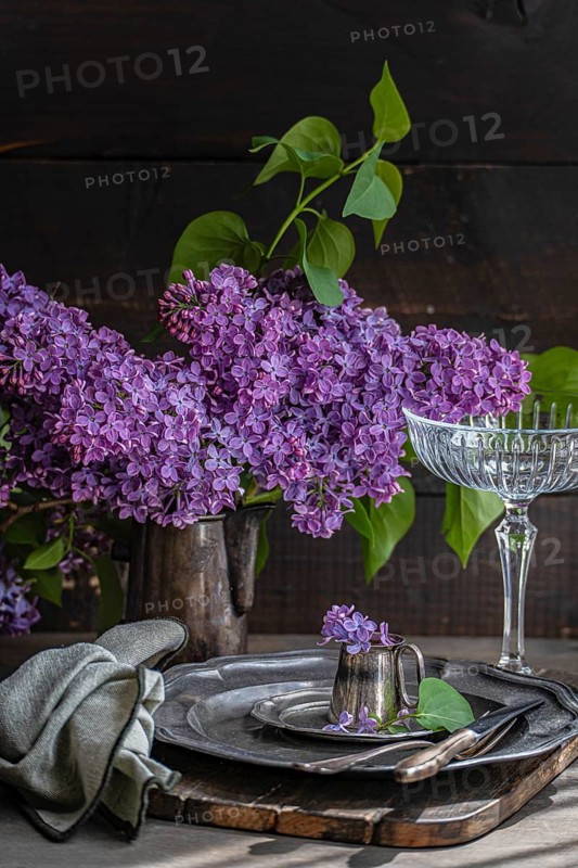 An elegant table setting with vibrant lilac blooms in a rustic pitcher. Vintage silverware and glassware complement the floral arrangement on a wooden table