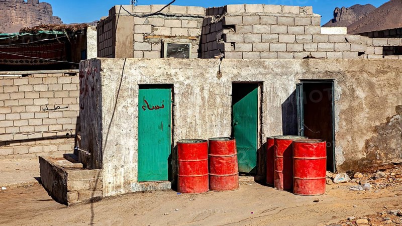 Simple huts with green doors in an arid desert landscape. Red barrels stand in front of the building, The public toilet in the Sahara in Algeria