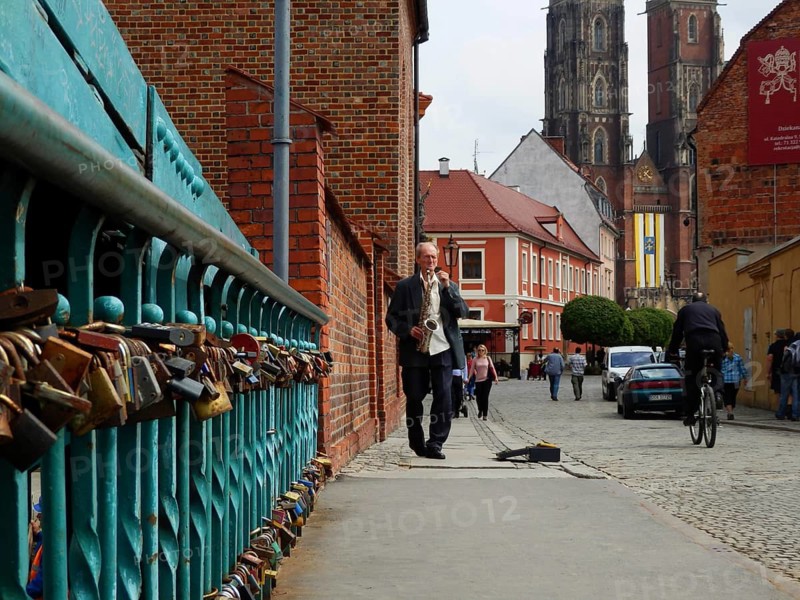 Scenic city view with cathedral bridge and love locks, musician with saxophone on sidewalk next to historic buildings, Wroclaw, Poland