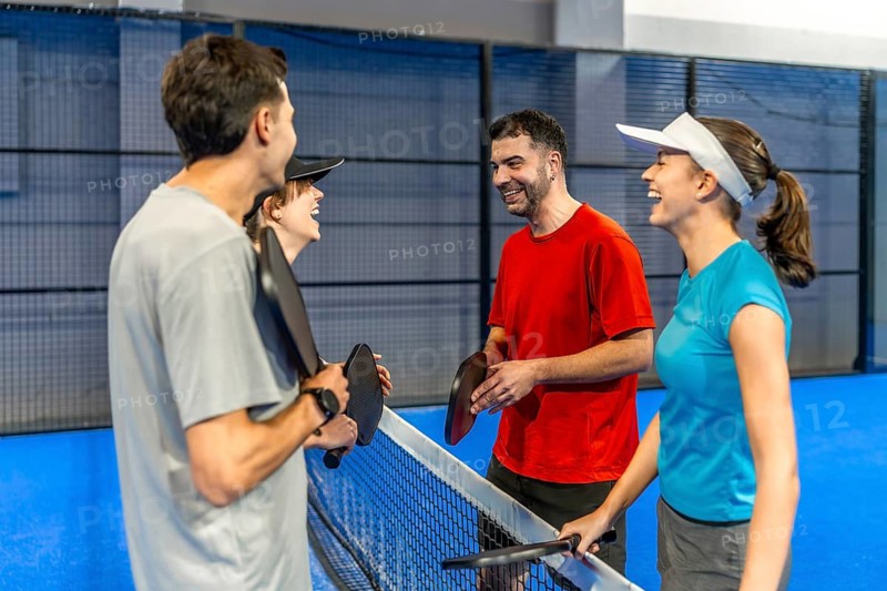Four friends laugh and chat on an indoor pickleball court, holding paddles after a game, enjoying casual conversation, camaraderie and active leisure together
