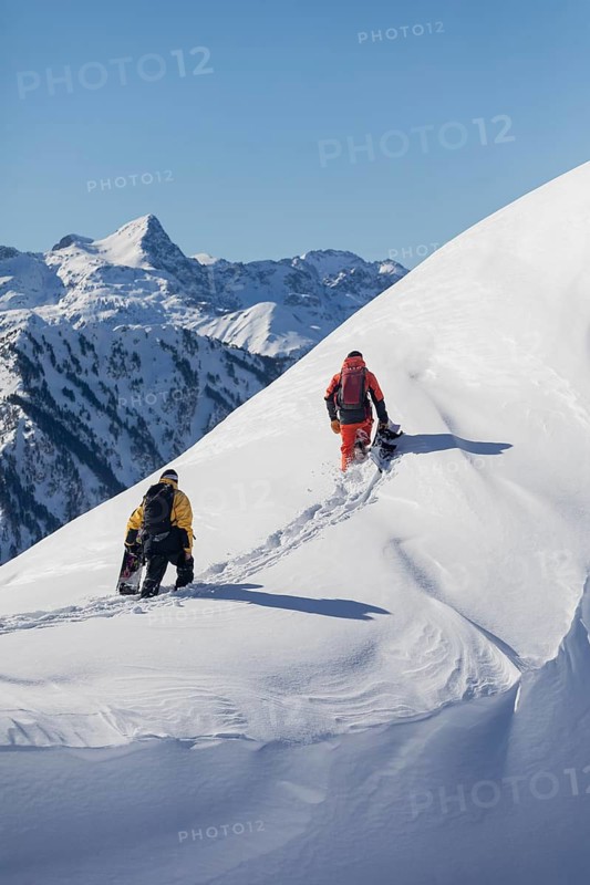 Two snowboarder friends trek up a snowy mountain slope under a clear blue sky. They carry snowboards, surrounded by vast snow covered peaks, embodying adventure and exploration
