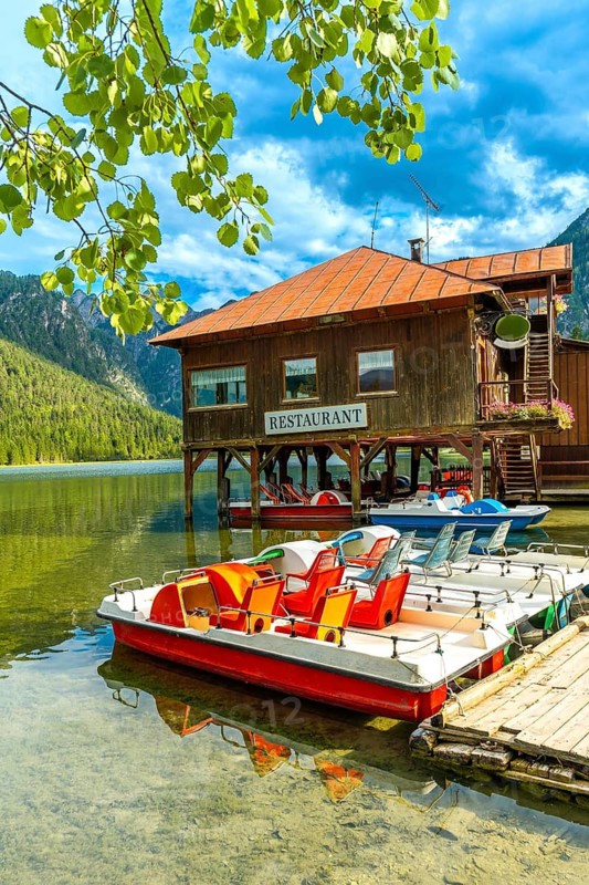 Colorful pedal boats sit docked below a wooden restaurant on lake dobbiaco with the dolomites rising in the background on a clear summer day