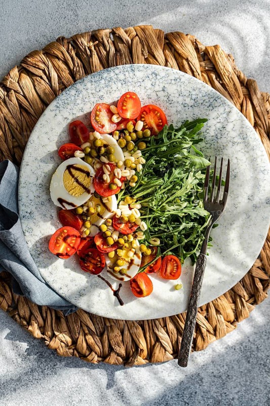 A wholesome keto lunch featuring boiled eggs, fresh rocket, cherry tomatoes, canned green peas, and sprouted green beans, topped with balsamic sauce on a decorative plate