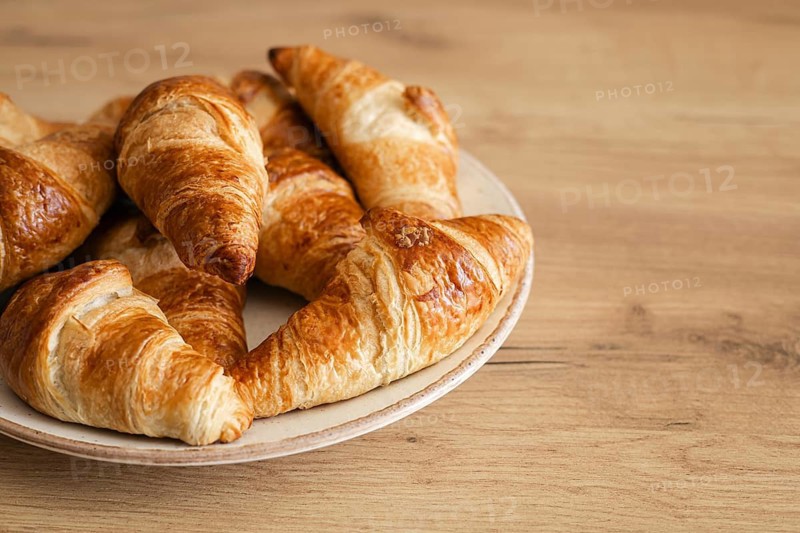 A plate of freshly baked homemade croissants sits invitingly on a wooden table. The golden, flaky pastry glistens, promising a delicious, buttery, and satisfying bite