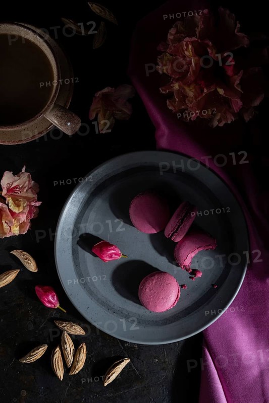 Purple macarons or macaroons cakes with cup of coffee on a black concrete background and pink textile. Hard light, low key. Top view, flat lay, close up