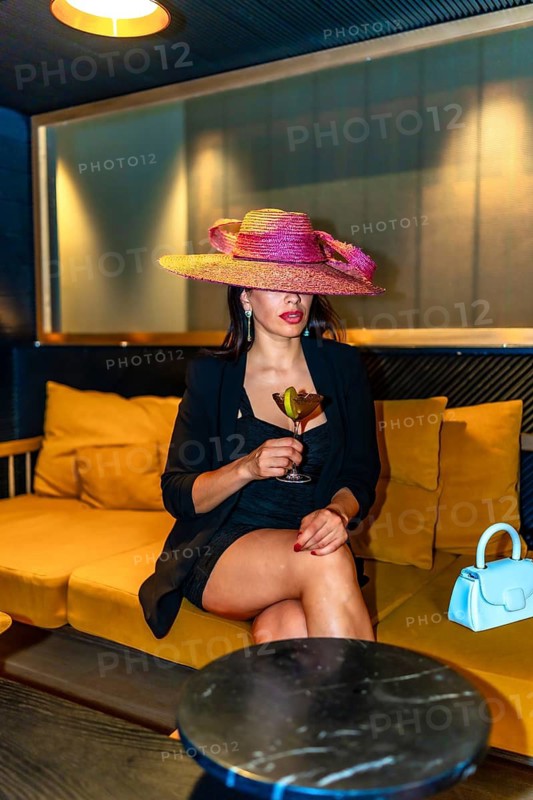 Vertical photo of a glamorous woman wearing dress and hat drinking a cocktail alone in a luxury pub