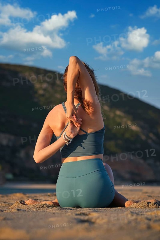 A woman sits in Gomukhasana, stretching in the peaceful embrace of a beach as the sun sets behind her and paints the sky
