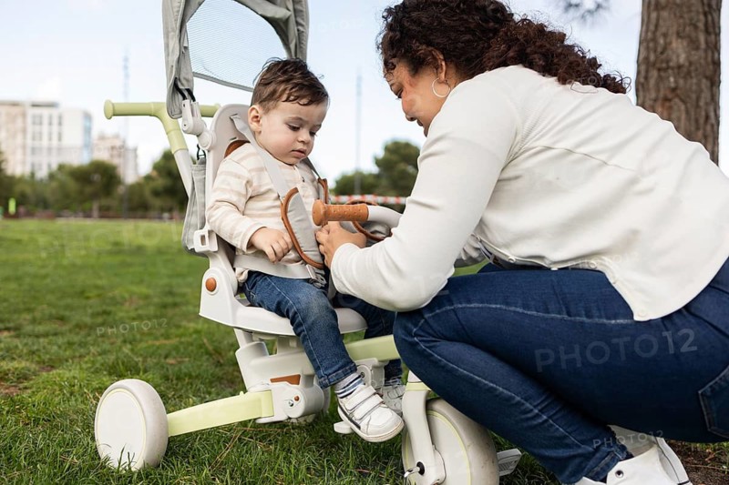A mother lovingly tends to her baby in a sprawling park. The baby is comfortably seated in a stroller, enjoying the warm afternoon. Trees and grass surround them, creating a serene setting