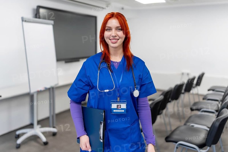 Young red haired medical student in blue scrubs and stethoscope stands in a bright modern lecture hall holding a folder, smiling confidently and ready for clinical training and career ahead