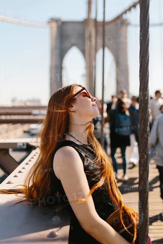 Young woman with sunglasses basks in the sun on a busy bridge in New York. Her long hair flows freely as she revels in the moment, surrounded by urban life and iconic architecture