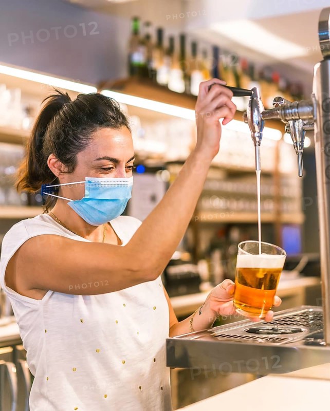 Photo session with a waitress with a face mask in a bar. New normality, security measures after the coronavirus pandemic, social distance, covid-19. Putting a glass with pole beer