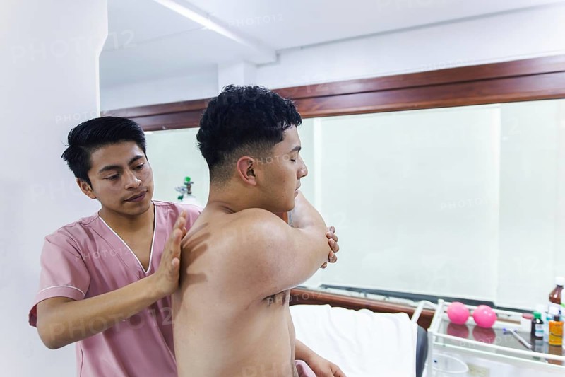 A physiotherapist aids a patient's shoulder movement in an Ecuador clinic. The setting includes therapy equipment, showcasing a professional health service environment