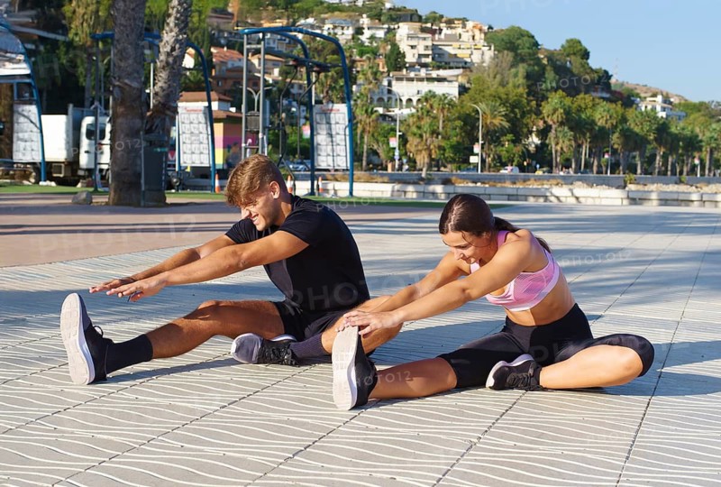 A woman and man engage in stretching exercises on a sunny seaside promenade, surrounded by palm trees and a scenic view of the ocean and distant hills
