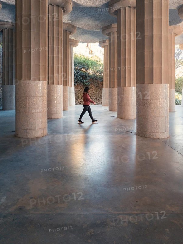 Side view of a woman striding across the smooth concrete floor under the towering stone columns of a famous architectural landmark in Barcelona