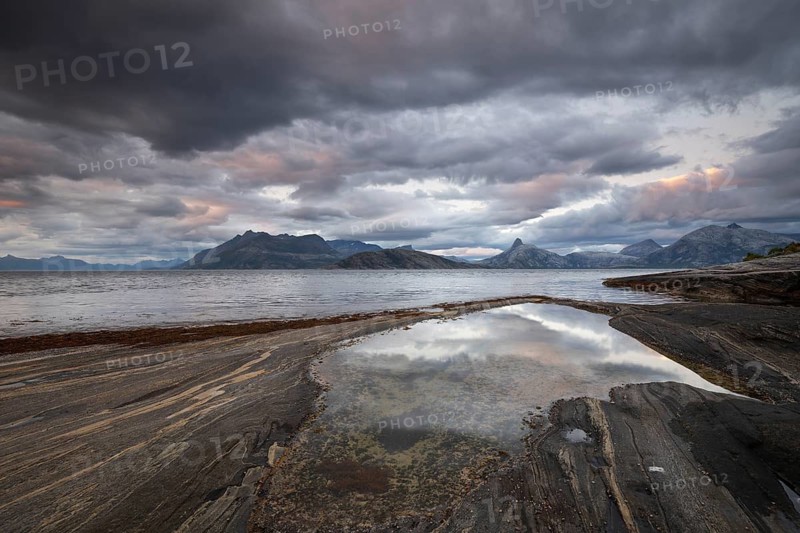 The Vestfjord in Nordland, Norway, offers a dramatic atmosphere near Bodø. Dark clouds are illuminated by the setting sun in shades of red and yellow. The rugged cliffs and mountains are reflected in the quiet fjord, which offers a breathtaking view of Lofoten