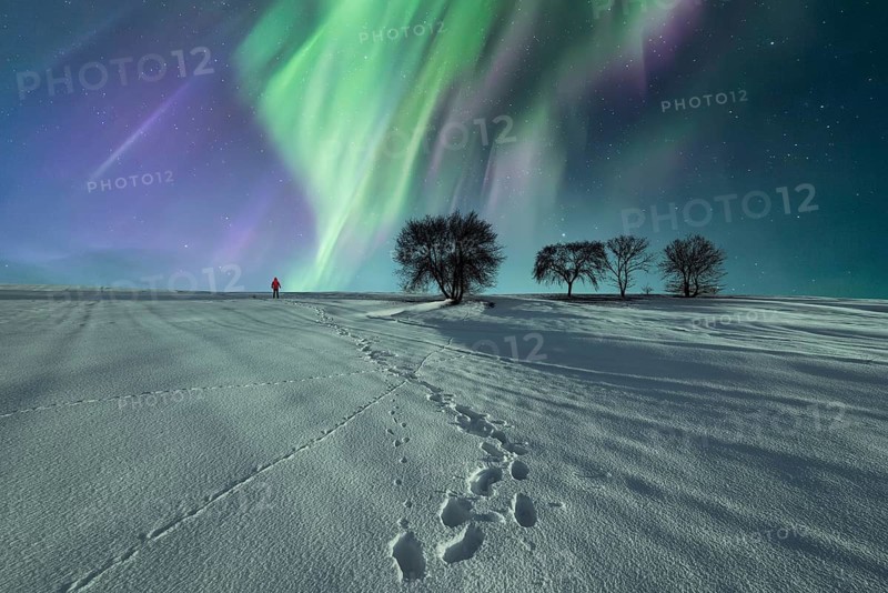 Unrecognizable person in red jacket walking through snowy terrain under a vibrant aurora borealis on a clear winter night, leaving footprints behind