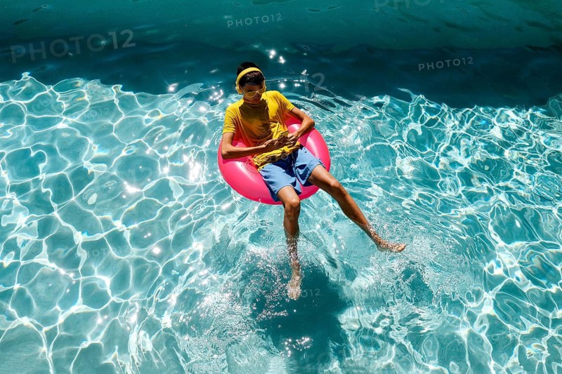 A person enjoys a sunny day, floating on a red inflatable ring in a clear blue pool. They wear a yellow shirt and sunglasses, embodying the essence of summer relaxation