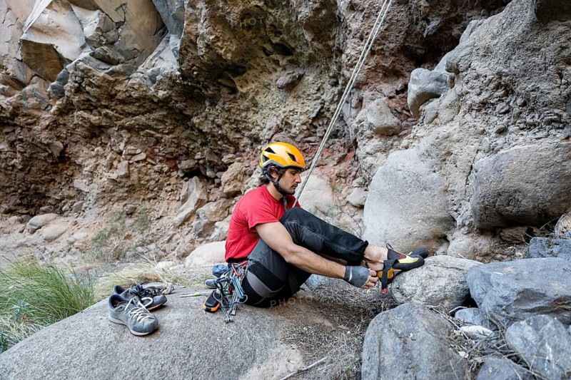 A climber sits on a rock, preparing for a traditional climb. Wearing a helmet and climbing gear, he adjusts his shoes against a rugged natural rock formation, surrounded by nature