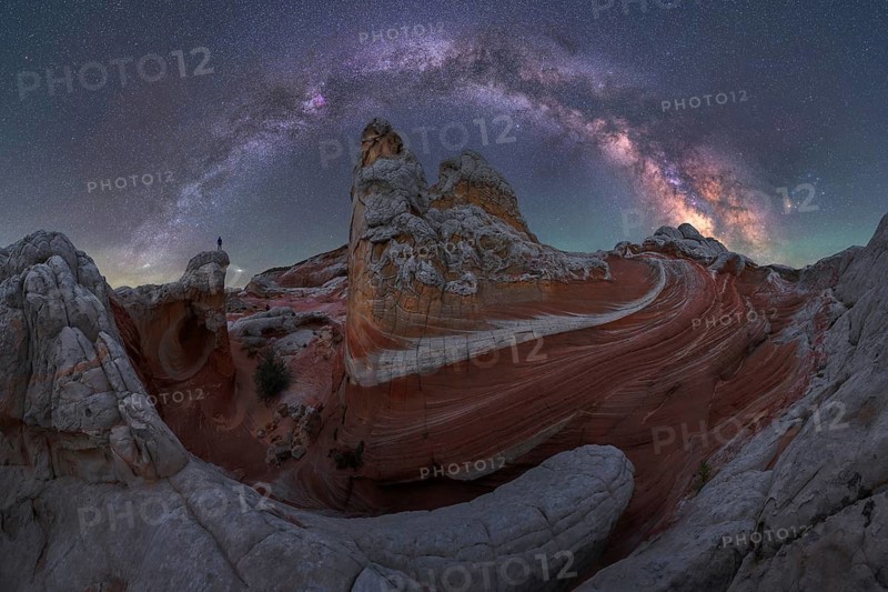 A man stands atop rock formations in White Pocket, Arizona, USA. The night sky is illuminated by the Milky Way, creating a stunning blend of natural beauty and cosmic wonder