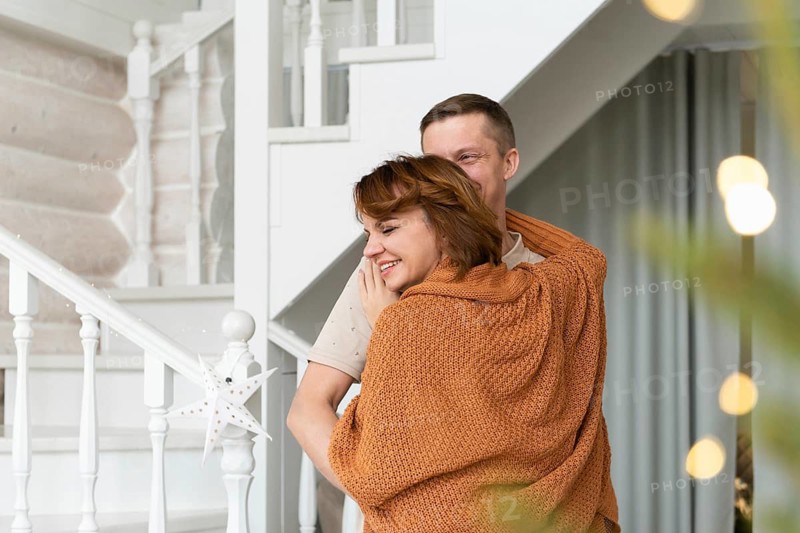 A man and a woman share a tender joyful embrace in a well-lit home, decorated with a subtle hint of holiday decor. Both are in casual attire, suggesting a warm, festive family occasion