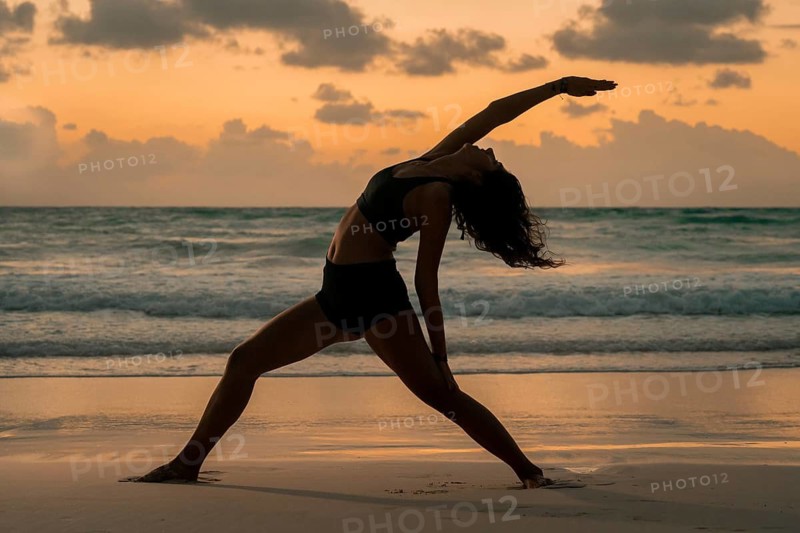 A woman performs a graceful yoga pose on a sandy beach, silhouetted against a stunning sunrise in Tulum, Mexico. The calm ocean waves and vibrant sky add a serene backdrop