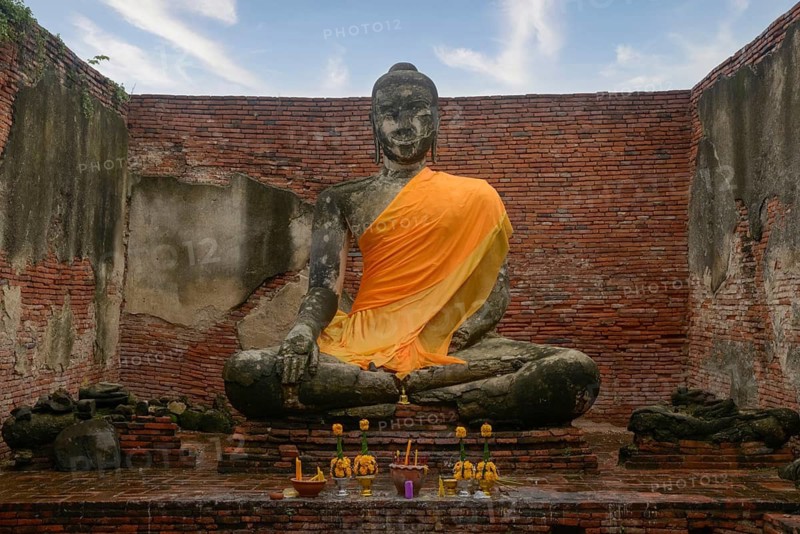 A Buddha statue adorned with an orange robe sits in the midst of ancient brick ruins of Ayutthaya. Yellow flowers and offerings are placed in front, under a partly cloudy sky in Chiang Mai, Thailand