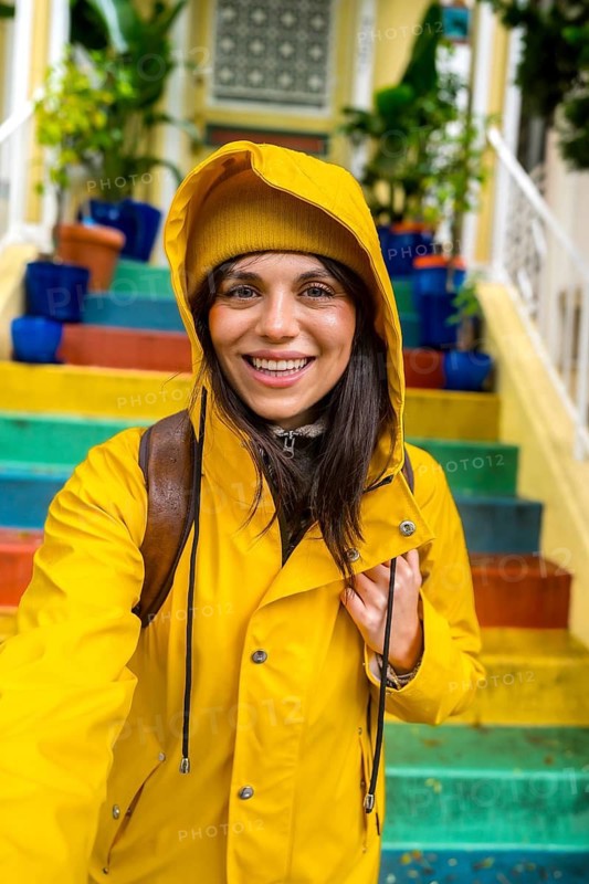 Woman smiling and enjoying urban travel, wearing a vibrant yellow rain jacket and beanie while standing on a flight of colorful rainbow stairs in san francisco