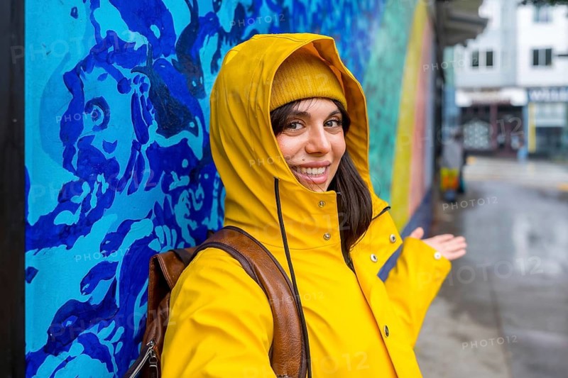 Young woman wearing a yellow raincoat and beanie smiling while standing by a colorful street art mural in an urban alley on a rainy day, showcasing travel and street style