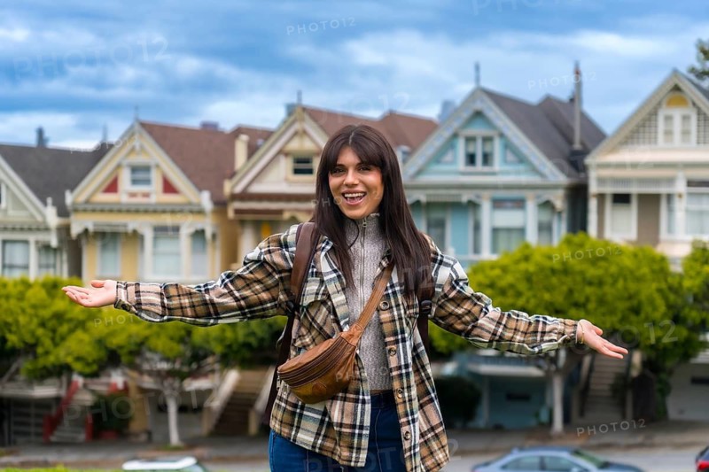 Young woman standing in front of the colorful victorian houses of alamo square park, smiling happily and spreading arms wide, expressing excitement and freedom while traveling