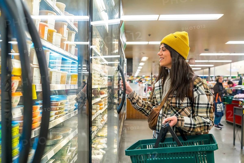 Woman holding a green shopping basket and opening a refrigerated display case to select a product, focusing on healthy eating and urban lifestyle during her grocery shopping