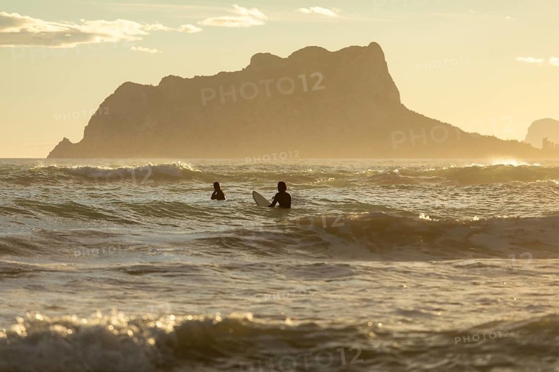 Two surfers enjoy the tranquil waves of the Mediterranean Sea at sunrise, with the Costa Blanca landscape creating a stunning backdrop as the sun casts a golden hue over the water