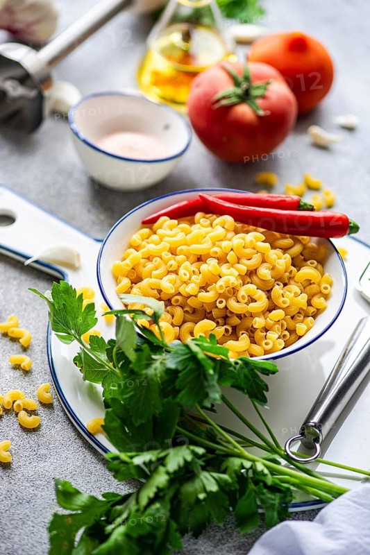 A vibrant image of uncooked chifferi rigati pasta in a white bowl, surrounded by fresh ingredients like chili peppers, parsley, tomatoes, and garlic on a rustic counter top