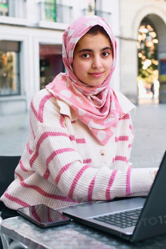 A arab student woman in a pink hijab and striped sweater uses a laptop at an outdoor cafe. Her smartphone rests nearby, embodying a blend of tradition and technology in a vibrant cityscape