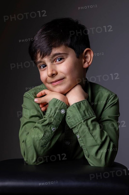 Young boy with dark hair and a content expression rests his chin on his hands, wearing a green shirt. The dark background highlights his cheerful and relaxed pose