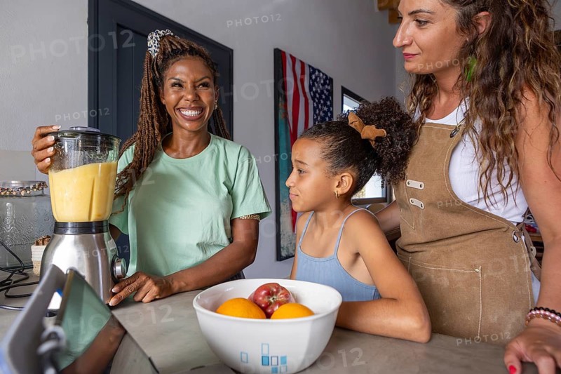 A joyful lesbian couple and their daughter enjoy family time in the kitchen, blending fruits. The scene captures love, pride, and everyday life in a diverse family