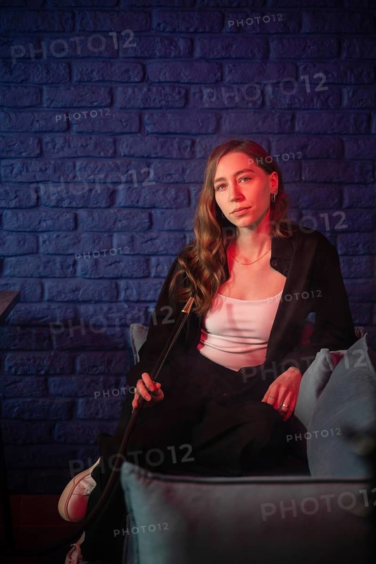 A woman sits in a dimly lit lounge, holding a hookah pipe against a blue brick wall. Red and blue lighting create a relaxed and modern ambiance
