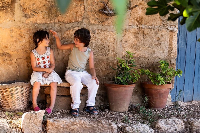 Two children sit on a stone bench near a rustic wall, with potted plants and a woven basket nearby. They are smiling and engaged in a playful moment outdoors