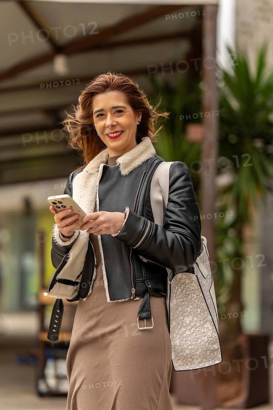 Happy businesswoman walking down a city street, using her smartphone and carrying a shopping bag