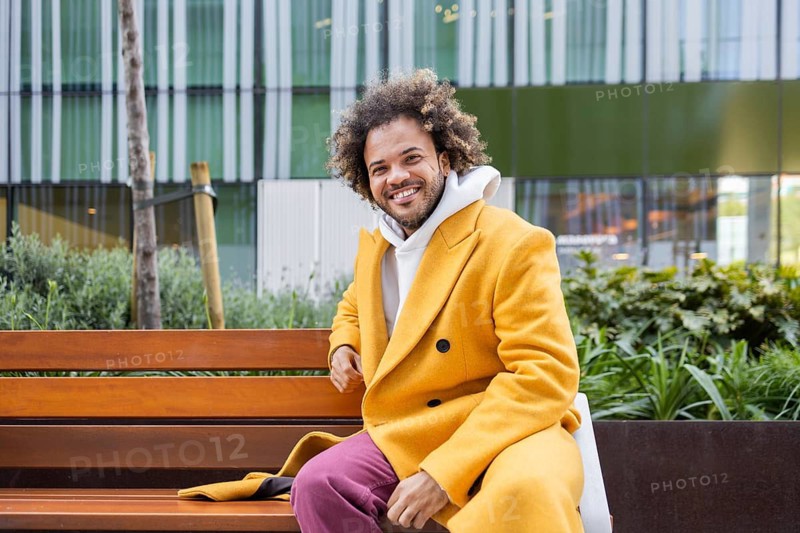 Afro man with curly hair in a bright yellow coat and pink pants sits on a wooden bench, smiling confidently with greenery and modern architecture in the background