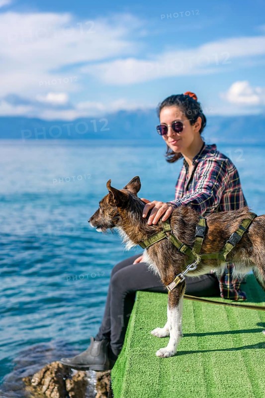 A woman, wearing sunglasses and a checked shirt, sits by the sea with her dog on a sunny day in Greece. The clear blue water and sky enhance the serene atmosphere